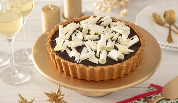 Foto da receita de Torta de Biscoito com Ganache de Café. Observa-se uma torta inteira sobre uma boleira dourada decorada com raspas de chocolate branco por cima e decorações de festa atrás.