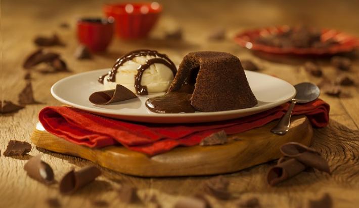 Fotografia em tons de marrom em uma bancada de madeira marrom. Ao centro, uma tábua de madeira, com um pano vermelho e um prato branco em cima contendo o petit gateau. Ao redor, há alguns pedaços de chocolates espalhados.