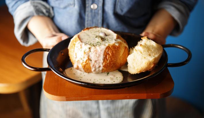 Fotografia de um chef segurando uma tábua de madeira, com uma panela grande preta em cima, com um pão aberto recheado com um creme de palmito que está transbordando e caindo na panela. Por cima, salsinha picada e salpicada.