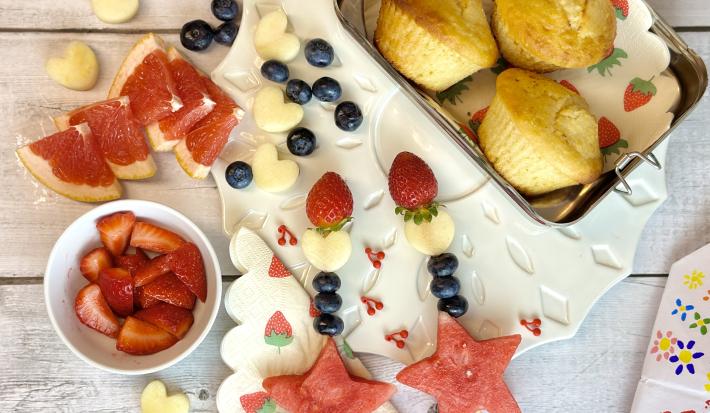 Fotografia em tons de vermelho em uma bancada de madeira clara, uma lancheira com muffin dentro, potinhos com frutas e brinquedos infantis.