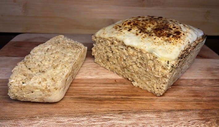 Fotografia de um pãozinho de aveia com queijo sobre uma tábua de madeira com uma fatia cortada.