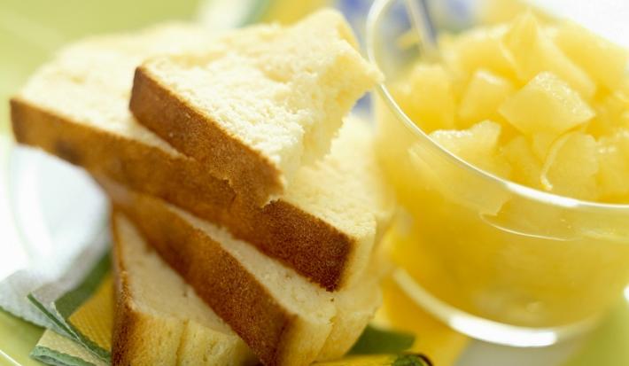 Fotografia em tons de amarelo, em uma bancada de madeira de cor branca. Ao centro, um prato verde contendo as fatias de pão com um recipiente ao lado contendo alguns pedaços de frutas.