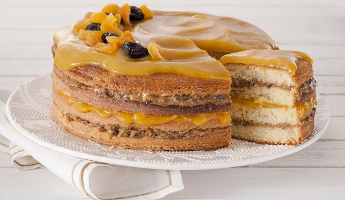 Fotografia em tons de amarelo em uma bancada de madeira branca com um pano branco com listras douradas e ao centro um prato branco com o bolo de coco com recheio de doce de leite com ameixa e geleia de damasco em cima.