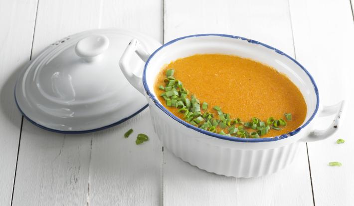 Fotografia em tons de branco, laranja e verde de uma bancada branca vista de cima. Ao centro um recipiente branco com alças e oval que contém um creme e ao lado a tampa do recipiente cor branca.