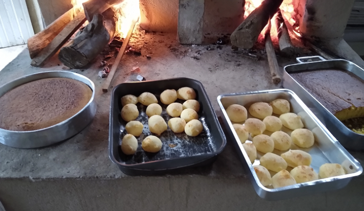 Foto em tons escuros da receita de pão de queijo servida em 30 porções divididas em duas formas sobre um fogão a lenha. Ao lado á mais duas formas com massas escuras e ao fundo estão dispostos diversos tocos de lenha