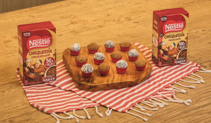Fotografia em tons de vermelho em uma bancada de madeira de cor marrom. Ao centro, um pano branco com listras vermelhas com uma tábua de madeira em cima contendo os brigadeiros. Ao lado, há duas caixinhas com cacau e chocolate em pó dois frades.