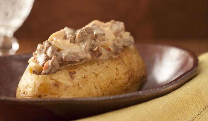 Fotografia em tons de marrom e pardo, em uma bancada contém um pano cor pardo com um garfo por cima, um recipiente marrom com uma batata com pedaços de carne com molho por cima e ao lado um copo transparente com agua