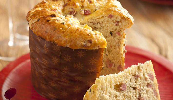 Fotografia em tons de vermelho e marrom, ao centro prato vermelho com um panetone fatiado, ao lado duas taças com bebida e uma decoração de natalina, ao fundo dois pratos empilhados, tudo sobre bancada de madeira rústica.