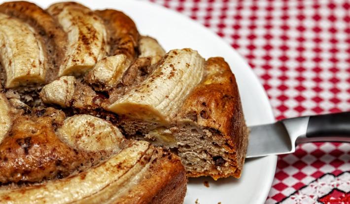 Fotografia em tons de amarelo, em uma bancada de madeira com uma toalha quadriculada vermelha. Ao centro, um prato redondo branco contendo o bolo de banana.