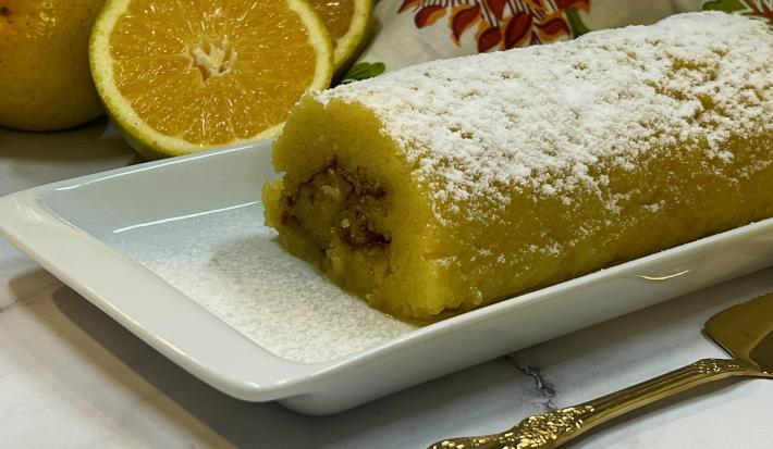 Foto da receita de Rocambole de Laranja. Observa-se um recipiente raso, retangular e branco contendo o rocambole polvilhado de açúcar de confeiteiro. Ao fundo, laranjas cortadas ao meio.