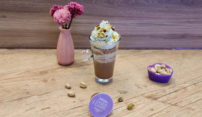 Fotografia em tons de lilás em uma bancada de madeira, com uma xícara de vidro e o Mochaccino Avelã com chantilly de pistache em cima para decorar. Em destaque, a cápsula de Dolce Gusto e ao fundo um vaso rosa com flores rosas.