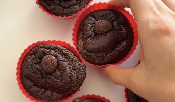 Fotografia mostra uma mão segurando um muffin de chocolate, fofinhos, com pedaço de chocolate derretido no topo, servidos em forminhas de silicone vermelha.
