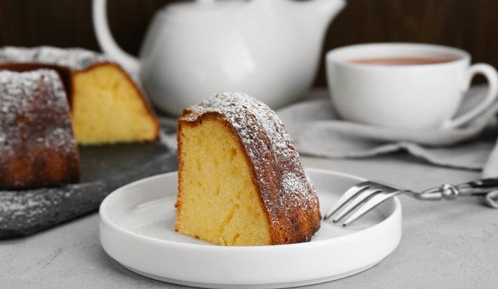 Fotografia de um pedaço de bolo em um prato branco, polvilhado com açúcar de confeiteiro, acompanhado por um garfo. Ao fundo, um bule e uma xícara de chá.