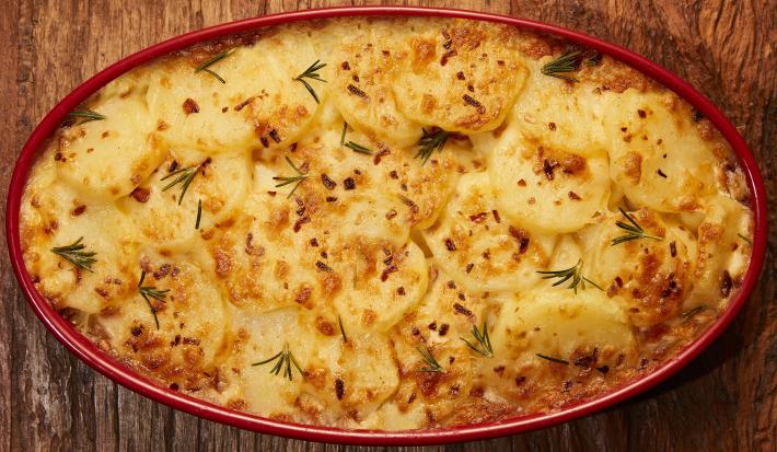 Foto bem aproximada de uma travessa vermelho dentro da qual está a receita de batatas gratinadas, decorada com ramos verdes de alecrim