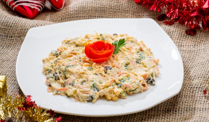 Fotografia de um salpicão tradicional cremoso com frango, cenoura, salsinha e uva passa em um prato branco quadrado. Por cima da salada, um tomate em formato de rosa e ao lado, uma folha de salsinha. Ao redor, decorações de Natal.