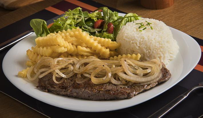 Fotografia em tons de marrom, branco e amarelo de uma bancada marrom, contém um prato redondo e branco com um bife com cebolas por cima, arroz, batatas fritas e salada como acompanhamento