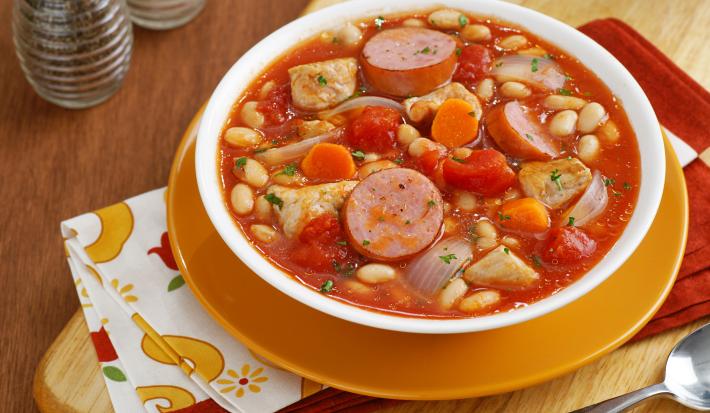 fotografia em tons de laranja e vermelho de uma bancada marrom vista de cima, contém um prato redondo laranja com um recipiente redondo branco que contém Cassoulet com pedaços de linguiça, feijão branco e abóbora.