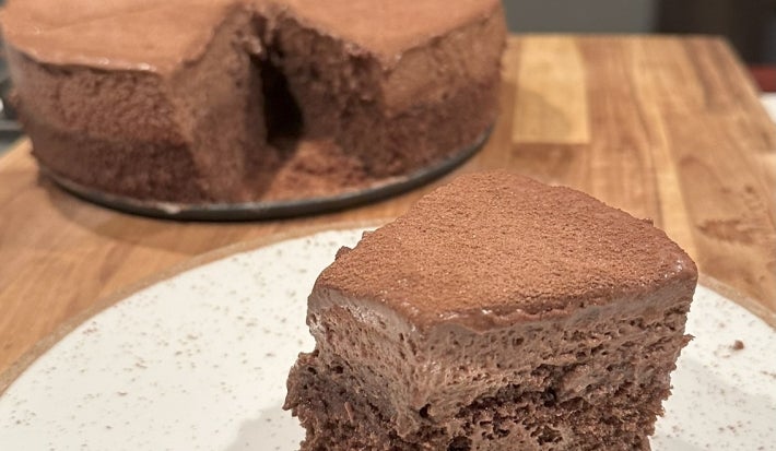 Fotografia de um bolo de chocolate caseiro, redondo e com uma cobertura de cacau. Uma fatia foi cortada, revelando seu interior úmido e fofo, apresentada em um prato branco, com um fundo de cozinha suave e acolhedor.
