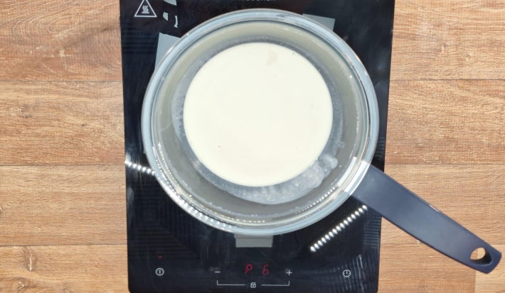 Creme de Leite cozinhando em banho maria sobre fogão portátil preto sobre bancada de madeira.