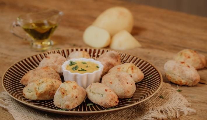 Fotografia em tons de marrom de uma bancada de madeira com um paninho de juta sobre ele um prato dourado redondo com croquetes de batata e ao centro um potinho redondo branco com molho. Ao fundo uma jarrinha com azeite e uma batata cortada.