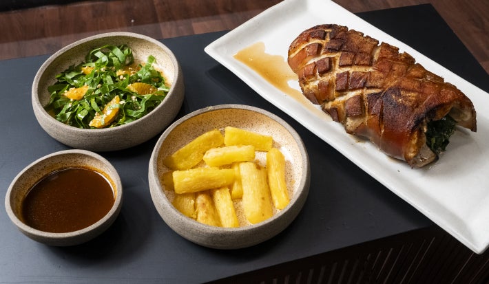 Pernil suíno assado com mandioca dourada, salada de folhas com laranja e molho servido à parte, apresentado em travessas.