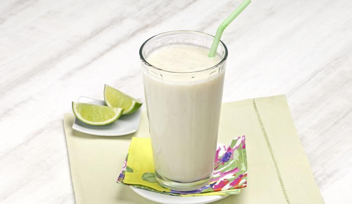 Fotografia em tons de verde em uma bancada de madeira cinza clara com um pano bege com detalhes verdes, um pratinho branco pequeno com um copo de vidro longo e grande com o smoothie de pera com limão. Ao lado, duas fatias de limão.