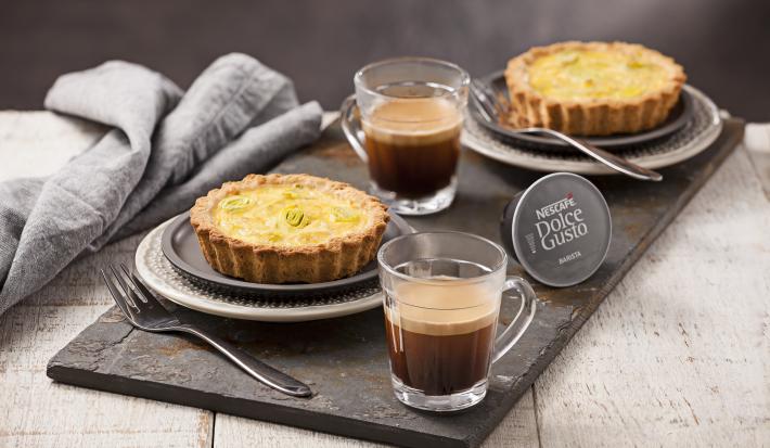 Fotografia em tons de cinza e dourado, ao centro dois copos com café, duas quiches assadas sobre  prato cinza e outro branco com borda decorada, com garfos ao lado, cápsula de café, sobre tábua grafite, guardanapo cinza, tudo sobre bancada em pátina.