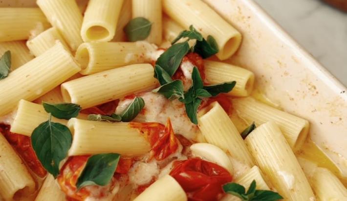 Fotografia em tons claros de uma travessa média, branca e retangular sobre uma mesa de mármore branca. Dentro da travessa tem uma receita de macarrão com tomate assado, manjericão, alhos inteiros assados e queijo.