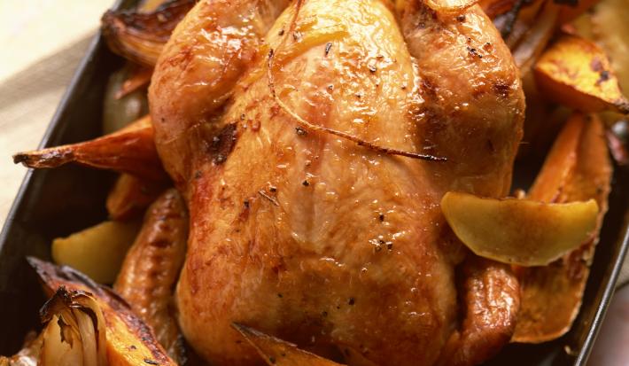 Fotografia vista de cima de um frango assado com batatas cortadas em meia lua, também assadas dentro de uma forma de vidro na cor preto. O assado está sobre um pano de mesa branco.