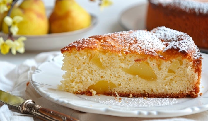 Fotografia de uma fatia de bolo de maçã em um prato branco, com uma textura leve e pedaços de fruta visíveis. O bolo é polvilhado com açúcar de confeiteiro.