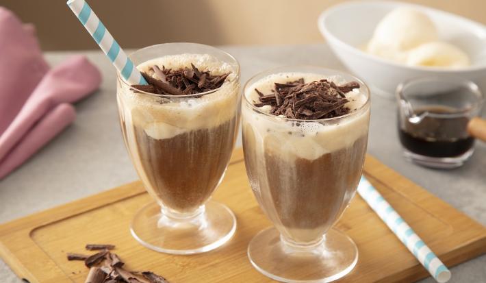 Foto em tons de marrom da receita de affogato de nescafé dolce gusto servida em duas xícaras de vidro dispostas sobre uma tábua de madeira com raspas de chocolate ao canto, além de canudos listrados em azul. Ao fundo uma xícara de café e sorvete.