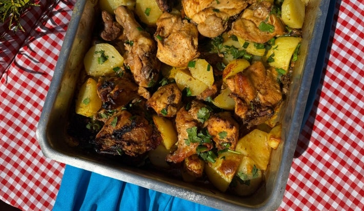 Frango assado dourado com batatas em rodelas levemente crocantes nas bordas, tudo coberto com cebolas caramelizadas e cheiro-verde fresco. Servido em assadeira rústica sobre mesa de madeira.