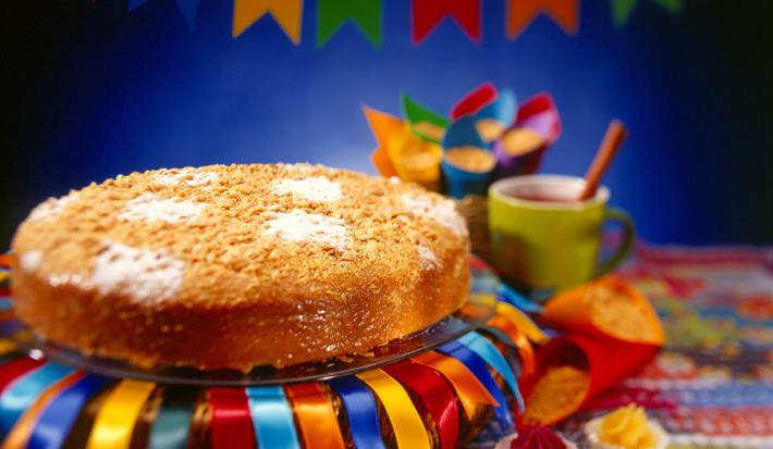 Fotografia em tons coloridos em uma bancada com uma toalha junina, um bolo redondo em cima de um prato com fitas de cetim decorando ele e bandeirolas de festa junina ao fundo.