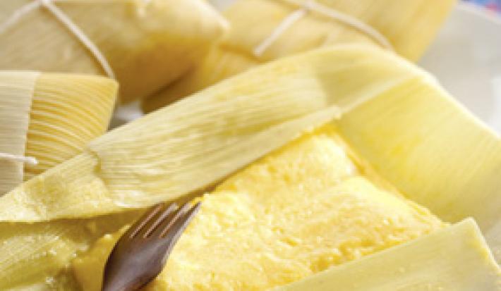 Fotografia em tons de amarelo em uma bancada de madeira, uma toalha colorida florida e algumas pamonhas de queijo coalho e alecrim em cima da bancada.