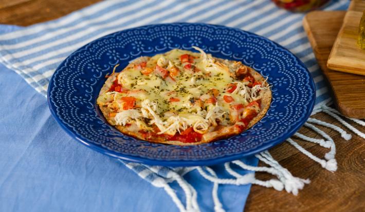 Fotografia em tons de azul com um prato azul ao centro. Em cima do prato existe uma receita de pizza de frigideira feita com frango desfiado, molho de tomate, queijo muçarela e orégano.