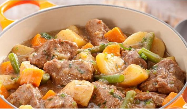 Fotografia em tons de laranja de uma bancada de madeira, com uma tábua de madeira, um pano quadriculado amarelo, vermelho e azul, uma colher prateada e uma panela laranja com alças com pedaços de carne, batata, cenoura e vagem.