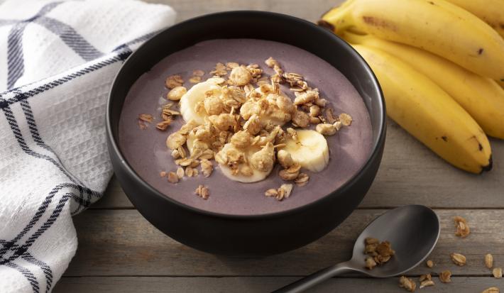 Fotografia em tons de branco e preto de uma bancada branca, sobre ela um recipiente redondo preto com açaí, um cacho de banana, uma colher e um paninho branco com listras pretas.