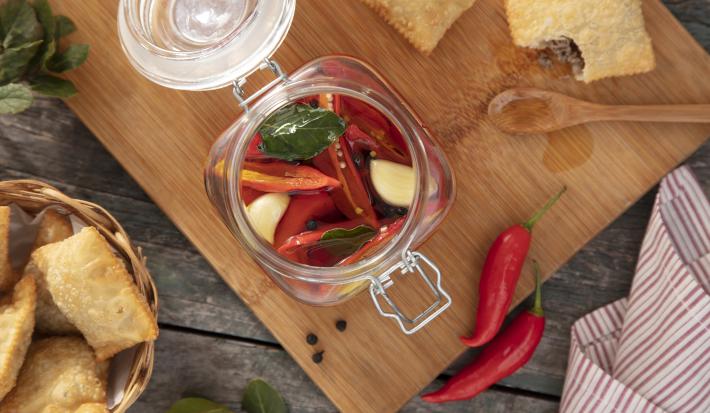 Imagem vista de cima de um pote de vidro de conserva de pimenta sobre uma tábua de madeira, com pastéis ao lado e algumas pimentas vermelhas