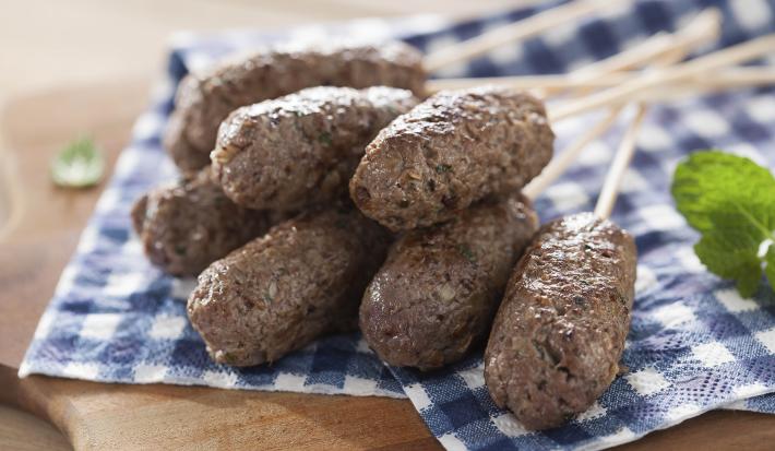 Fotografia em tons de azul em uma bancada de madeira com um pano xadrez azul e mini kaftas em palitos em cima dele. Ao lado, folhas de hortelã.