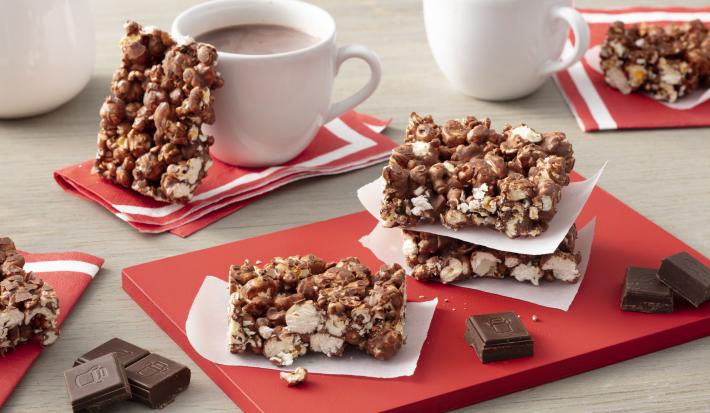 Fotografia em tons de vermelho em uma bancada de madeira clara com barras de pipoca com chocolate espalhadas pela mesa e em cima de uma tábua vermelha. Ao fundo, duas xícaras de chocolate quente com uma barra de pipoca com chocolate apoiada em uma delas. Fotografia em tons de vermelho em uma bancada de madeira clara com barras de pipoca com chocolate espalhadas pela mesa e em cima de uma tábua vermelha. Ao fundo, duas xícaras de chocolate quente com uma barra de pipoca com chocolate apoiada em uma delas.