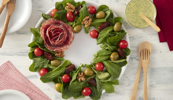 Foto da receita de Salada Guirlanda de Natal. Observa-se a rúcula, os tomates, a azeitona e as tâmaras em formato de guirlanda no prato branco, enquanto o salame está em formato de rosa ao lado esquerdo.