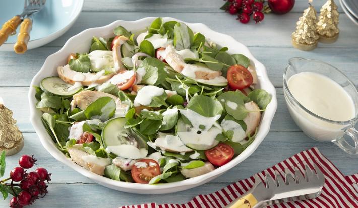 Foto em tons claros da receita de salada de chester com molho de iogurte servida em um bowl grande de porcelana branca com uma xícara de iogurte ao lado sobre uma mesa de madeira azul