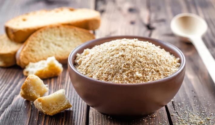 Fotografia em tons claros de um recipiente pequeno e fundo marrom sobre uma mesa de madeira escura. Ao lado do recipiente tem alguns pedaços de pão, e no fundo tem três fatias de pão.