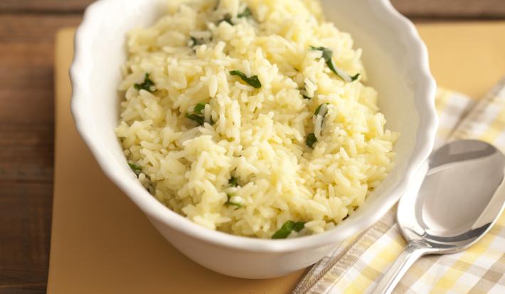 Fotografia em tons de amarelo em uma bancada de madeira com um pano listrado em em bege e amarelo e um prato fundo branco com o arroz com rúcula.