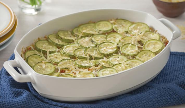 Fotografia em tons de verde em uma mesa de madeira com uma toalha branca, um pano azul, um refratário branco oval com a abobrinha gratinada dentro dele.