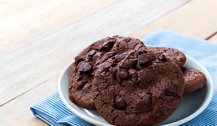 Fotografia de quatro cookies de chocolate com gotas de chocolate dentro de um recipiente de vidro branco um pouco fundo. O recipiente está por cima de um paninho azul com listras brancas sobre uma mesa de madeira.