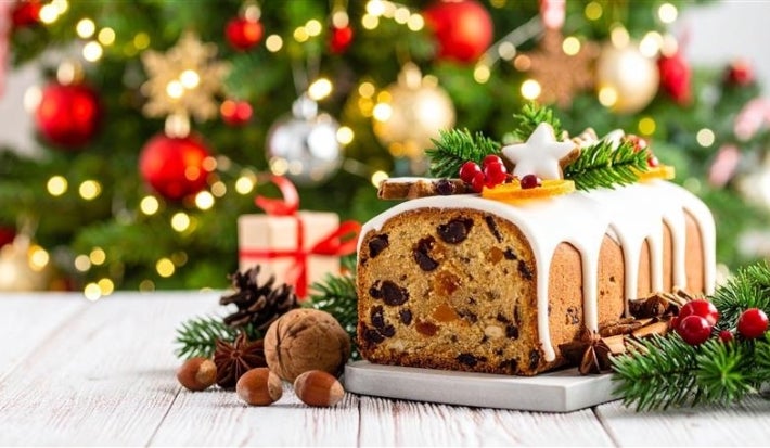 Fotografia de um bolo de frutas natalino, coberto com glacê branco e enfeitado com um biscoito em forma de estrela, frutas secas e ramos de pinheiro. Ao fundo, uma árvore de Natal iluminada e presentes.