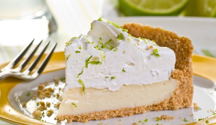 Fotografia de uma fatia generosa de torta de limão com raspas e chantilly sobre um prato de vidro com o fundo branco e a borda amarela.