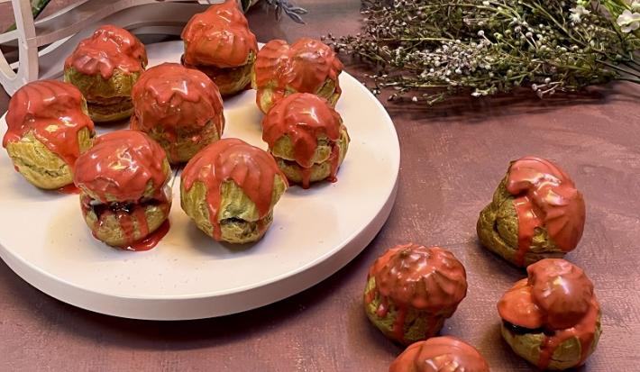 Foto em tons de vermelho da receitas de carolinas de frutas vermelhas servidas em diversas porções, algumas sobre uma base redonda branca e outras sobre a mesa vermelha. Ao fundo há flores para decoração