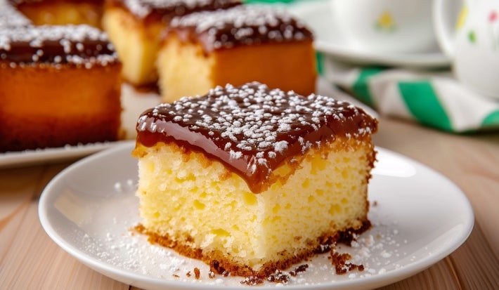 Fotografia de um pedaço de bolo quadrado com crosta dourada e interior amarelo, coberto com uma camada de chocolate brilhante e polvilhado com açúcar de confeiteiro.
